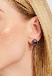 Kolju kujulised stud-kõrvarõngad, millel on lilla läige ja mustad vääriskivide aksessuaarid, valmistatud metallist ja tekstureeritud pinnaga.