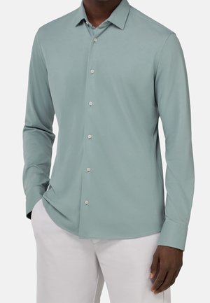 Mężczyzna ubrany w jasnozieloną koszulę na długi rękaw z białymi guzikami oraz jasne spodnie, stojący z jedną ręką w kieszeni.