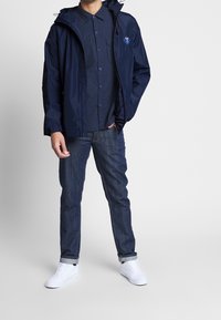 Granatowa wodoodporna kurtka z kapturem, z logo na piersi, na ciemnej koszuli na guziki. Noszona z ciemnymi dżinsami i białymi trampkami.
