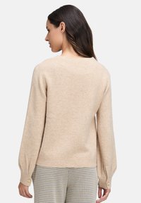Frau mit langen dunklen Haaren, die einen beigen Strickpullover und karierte Hosen trägt, von hinten vor einem weißen Hintergrund gezeigt.