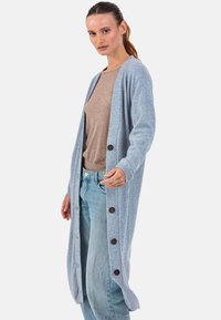 Cardigan long bleu clair en tissu doux, avec des boutons sur le devant, coupe décontractée et poignets côtelés, porté sur un haut beige.