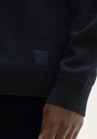 Granatowy sweter robiony na drutach z prążkowanymi mankietami i dołem, z teksturowaną etykietą w lewym dolnym rogu. Materiał wydaje się miękki i elastyczny.