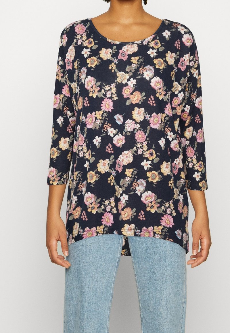 Marinblus med blommönster och trekvartslånga ärmar, med rosa och gula blommor, rund halsringning och avslappnad passform, kombinerad med ljusblå jeans.