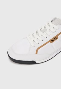 Weißer Sportschuh mit einem strukturierten Canvas-Obermaterial, ausgestattet mit einem goldenen Akzentstreifen und einer verstärkten schwarzen Gummisohle. Die Schnürsenkel sind weiß.