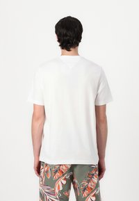 T-shirt en coton blanc avec un col rond et des manches courtes, présentant une texture subtile. Porté avec un short vert à motifs.