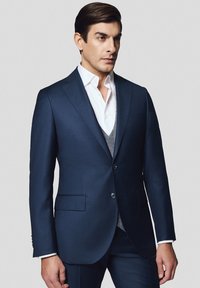 Granatowa marynarka z dwiema guzikami, klapami w stylu notch i bocznymi kieszeniami, noszona na białej koszuli i szarym kamizelce o teksturowanym wzorze.