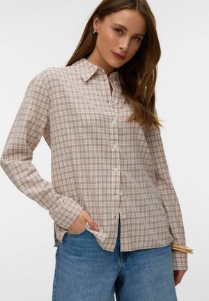 Žena nosiaca bežovú a bielu károvanú košeľu na zapínanie a modré džínsy, s jednou rukou v vrecku a v druhej drevené paličky na jedlo.