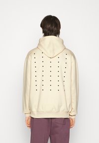Felpa beige con cappuccio, dotata di aperture circolari nere sulla schiena, polsini e orlo a costine, con una vestibilità rilassata e un texture morbida.