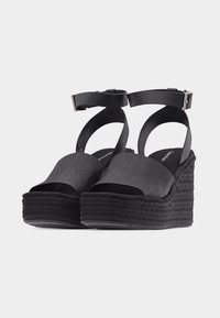 Sandales compensées noires avec brides à la cheville et semelles plateformes texturées. Design à bout ouvert avec larges brides avant présentant des motifs de couture subtils.