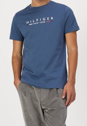 Niebieska bawełniana koszulka z logo "HILFIGER NEW YORK" w białym i czerwonym kolorze. Krótkie rękawy, okrągły dekolt i luźny krój. W zestawie z szarymi spodniami z sztruksu.