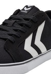 Czarne płócienne trampki z białą gumową podeszwą, czarnymi sznurówkami i wyraźnymi białymi akcentami w kształcie chevronu po bokach. Logo marki na języku.