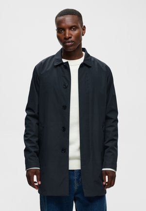 Homme portant un manteau noir boutonné par-dessus un pull blanc et un jean bleu, debout devant un fond uni gris clair.