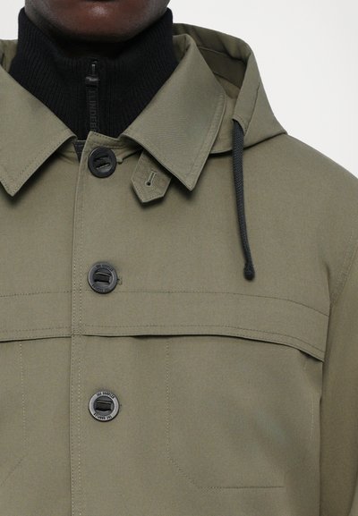 Olivgrüne Jacke mit hohem Kragen und Kapuze, ausgestattet mit vier schwarzen Knöpfen, einem gefütterten Nacken und kontrastierendem Rippstrick-Muster.