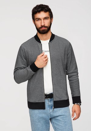 Homme aux cheveux foncés et à la barbe, portant une veste grise zippée avec des poignets et un ourlet noirs, une chemise blanche, un jean bleu clair et une montre-bracelet.