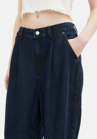 Ciemnoniebieskie, wysokie dżinsy z plisami, bocznymi kieszeniami i metalowym zapięciem. Gładka tekstura i prosty krój nogawek.
