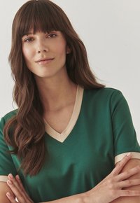 Green v-neck top with beige trim at the neckline and sleeves. Soft fabric with a smooth texture, fitted design, and short sleeves.