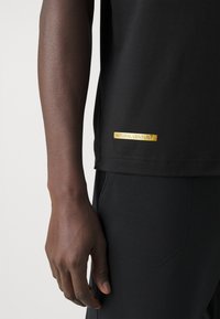 Rechterarm en hand van een persoon in een zwart shirt en zwarte broek, de zoom van het zwarte shirt toont een klein gouden label met de tekst "NATURAL VENTUS".