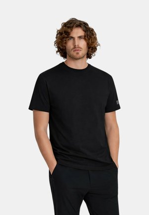 Uomo con capelli ricci che indossa una maglietta nera semplice a maniche corte e pantaloni neri, in piedi con le mani in tasca davanti a uno sfondo bianco.