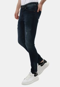 Donkerblauwe skinny jeans met verwoestte details aan de voorkant, gecombineerd met witte en zwarte sneakers. Gladde denimtextuur, eigentijds ontwerp.