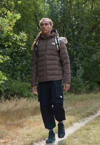 Rudas pūkuotas džemperis, juodi krovinių šortai, juodi žygio batai. Lauko aplinka su žalia augmenija, vaikštoma žemės taku. Matomi tekstūruoti medžiagų paviršiai.