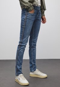 Henkilö, jolla on yllään siniset slim fit -farkut, joissa on sivuviivat, sekä valkoiset tennarit, seisoo harmaalla lattialla yksinkertaista seinää vasten.