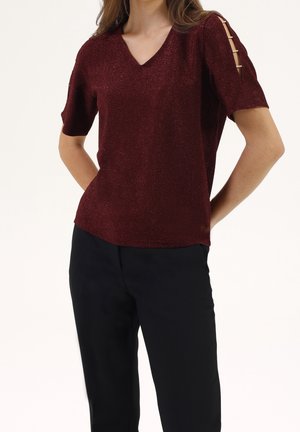 Kobieta w bordowej, błyszczącej bluzce z krótkim rękawem z wycięciami na rękawie w formie drabinki oraz czarnych spodniach, stojąca z rękami za plecami.