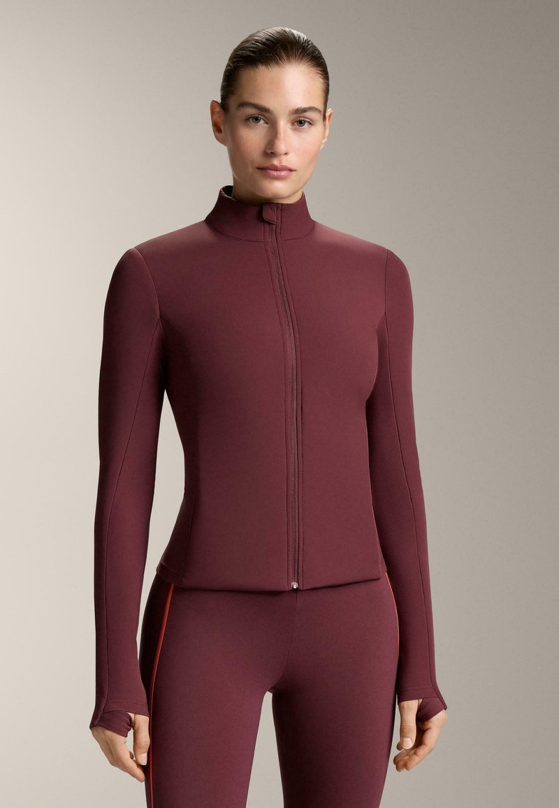 Atletska jakna boje burgundca sa patentnim zatvaračem, dugim rukavima, visokim ovratnikom i otvorima za palce. Glatka tekstura, kroj prilagođen tijelu i minimalni šavovi.