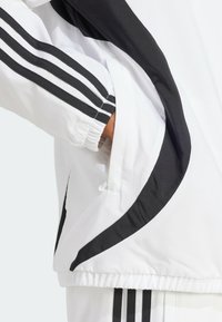 Mano en el bolsillo de una chaqueta blanca con rayas negras y pantalones blancos a juego con rayas negras, mostrando puños elásticos y diseño deportivo.