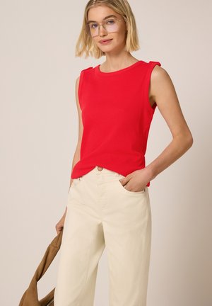 Femme aux cheveux blonds courts portant des lunettes transparentes, haut rouge sans manches, pantalon crème, tenant une veste marron dans la main gauche.