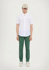 Homem a usar um gorro bege, camisa de botão de mangas curtas branca, calças verdes e sapatilhas brancas, de pé contra um fundo simples.