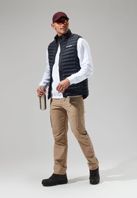 Zwarte puffervest over een wit langemouwen shirt, khaki cargobroek en zwarte laarzen. Houdt een roestvrijstalen waterfles vast. Rode pet en zonnebril.