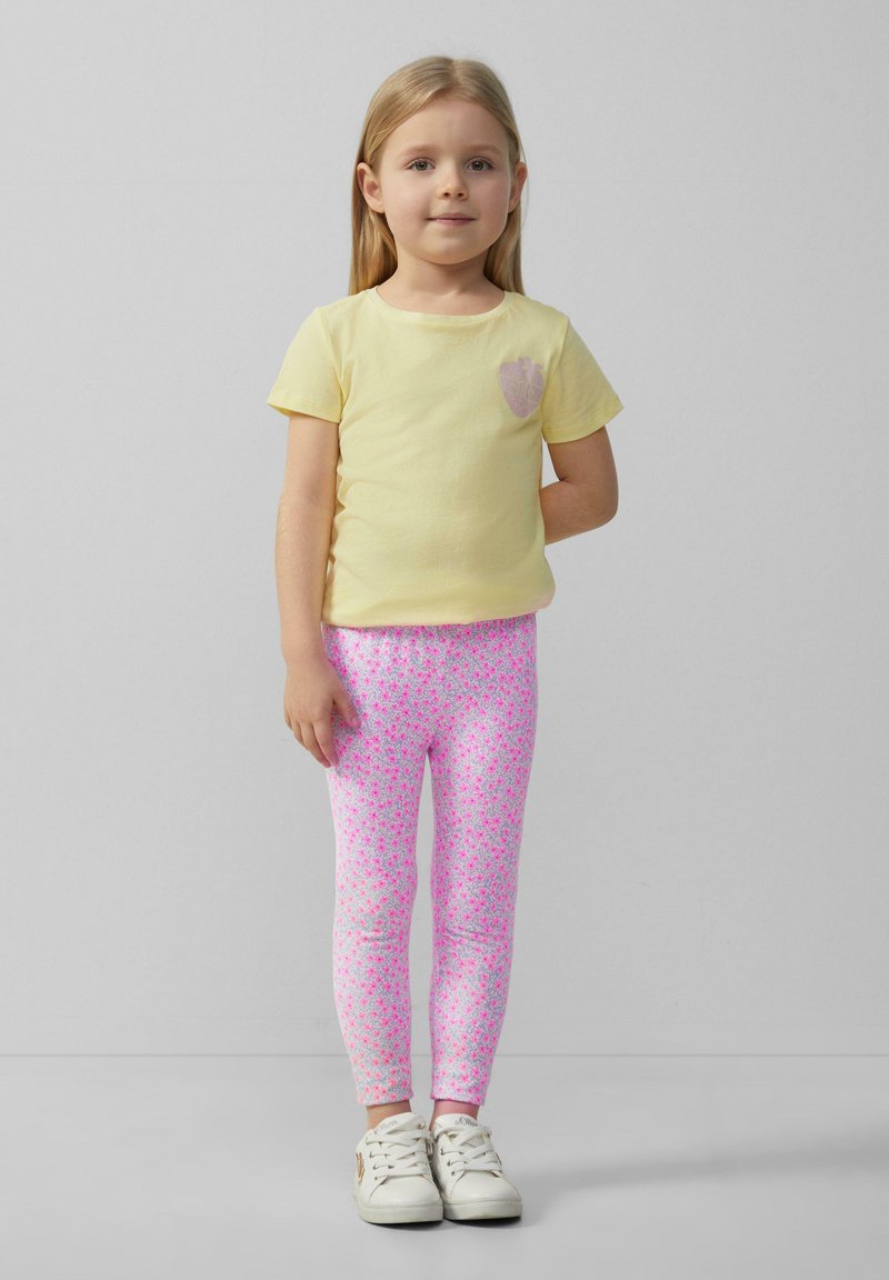 Geel katoenen T-shirt met een hartontwerp op de borst, gecombineerd met roze bloemenleggings. Witte sneakers maken de outfit compleet.