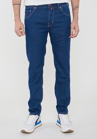 Jeans in denim blu con una vestibilità slim, cuciture rosse a contrasto, design a cinque tasche e chiusura con bottone. Abbinati a sneakers bianche e blu.