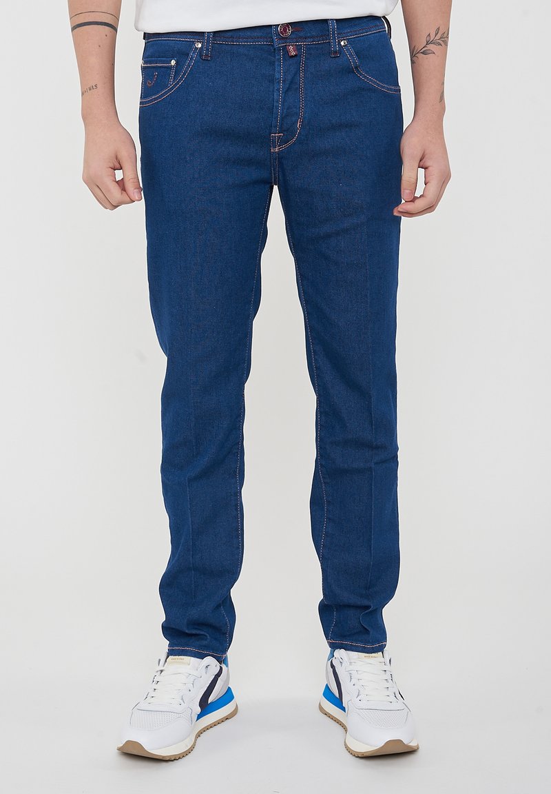 Jeans in denim blu con una vestibilità slim, cuciture rosse a contrasto, design a cinque tasche e chiusura con bottone. Abbinati a sneakers bianche e blu.
