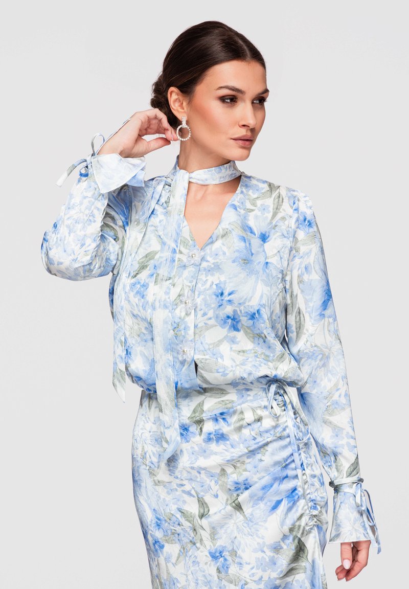 Mujer con cabello oscuro recogido en un moño bajo, vestida con una blusa de patrón floral azul claro y falda a juego, complementada con pendientes de aro y un lazo en el cuello.