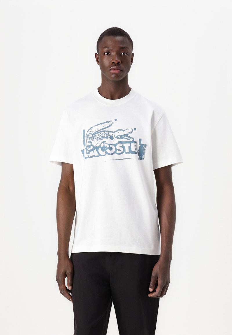 Camiseta de algodón blanca con un gráfico del logo de Lacoste en azul. Manga corta y cuello redondo clásico. Corte simple y recto.