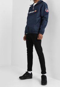 Marinblå vindjacka med ficka fram, logodetalj och justerbar dragsko i fållen, kombinerad med svarta slim fit-jeans och svarta sneakers.