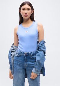 Jeune femme portant un top sans manches bleu clair et un jean, en train d’enlever partiellement une veste en jean, debout devant un fond blanc.