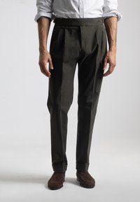 Pantalones de color verde oscuro con pliegues, que presentan una cinturilla ancha y un bolsillo lateral. Textura suave y ajuste entallado, combinados con zapatos de ante marrón.