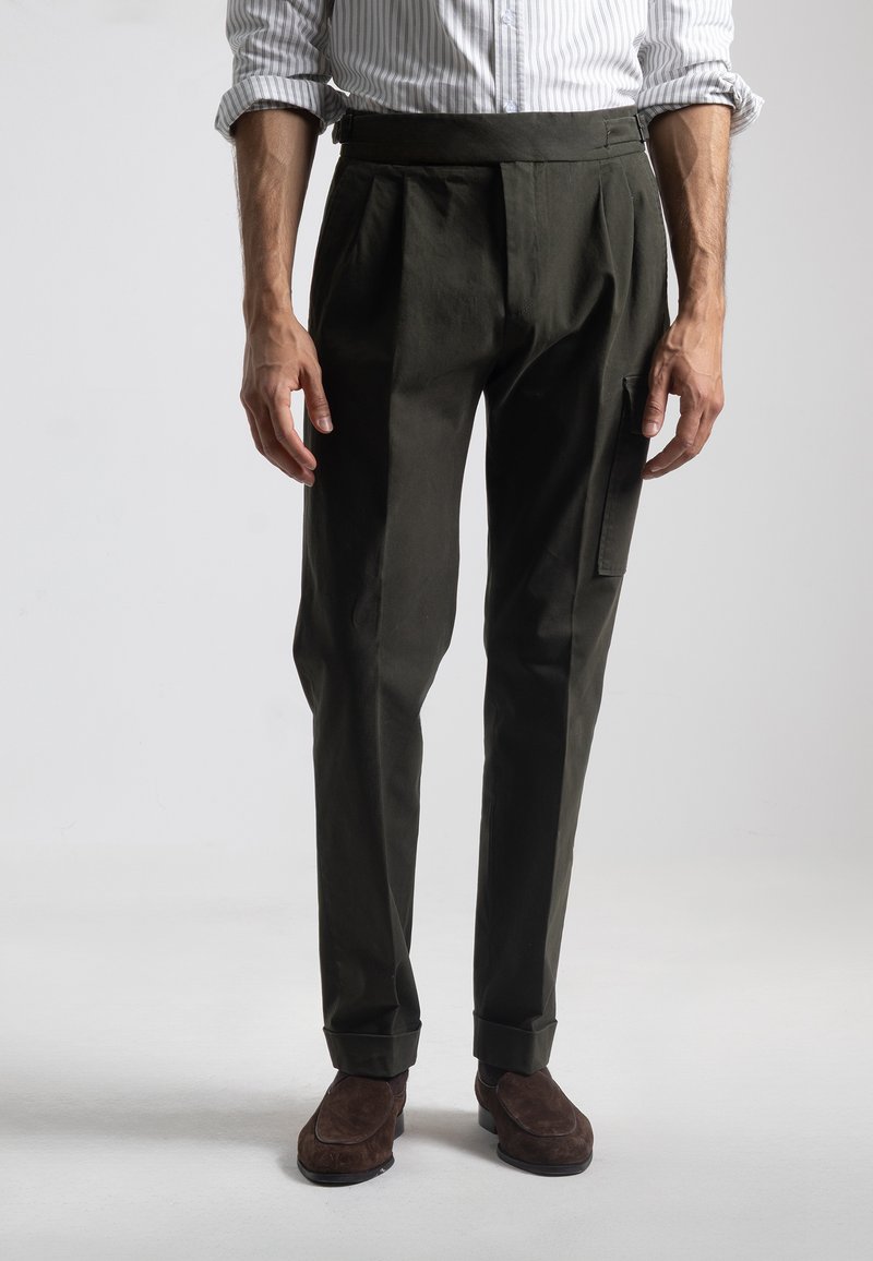 Pantalones de color verde oscuro con pliegues, que presentan una cinturilla ancha y un bolsillo lateral. Textura suave y ajuste entallado, combinados con zapatos de ante marrón.