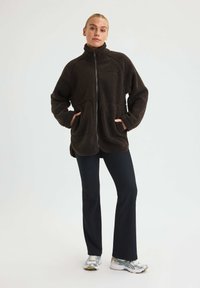 Nainen, joka on pukeutunut mustaan fleece-huppariin, kädet taskuissa, mustiin housuihin ja valkoisiin tennareihin, seisoo yksinkertaista taustaa vasten.