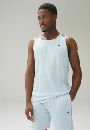 Mann trägt hellblaues ärmelloses Sportoberteil und passende Shorts, steht mit einer Hand in der Tasche vor einfarbigem Hintergrund.