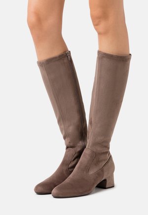 Knee-high taupe suede boots with side zipper and low block heel worn on bare legs against white background.