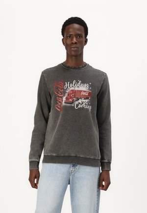 Grijze sweatshirt met lange mouwen, voorzien van een voorkant graphic van een Coca-Cola vrachtwagen en tekst. Blauwe jeans met een vervaagde uitstraling.