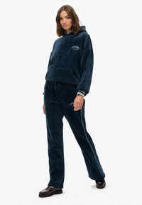 Navyblauer Velours-Trainingsanzug mit Kapuzensweatshirt und Fronttasche sowie passenden Hosen mit seitlichen Paspeln, akzentuiert durch weiße Etiketten.