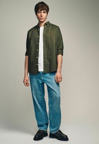 Chemise verte à boutons portée sur un t-shirt blanc, associée à un jean bleu et des chaussures noires. Le modèle a les cheveux courts et bouclés et se tient dans une pose neutre.