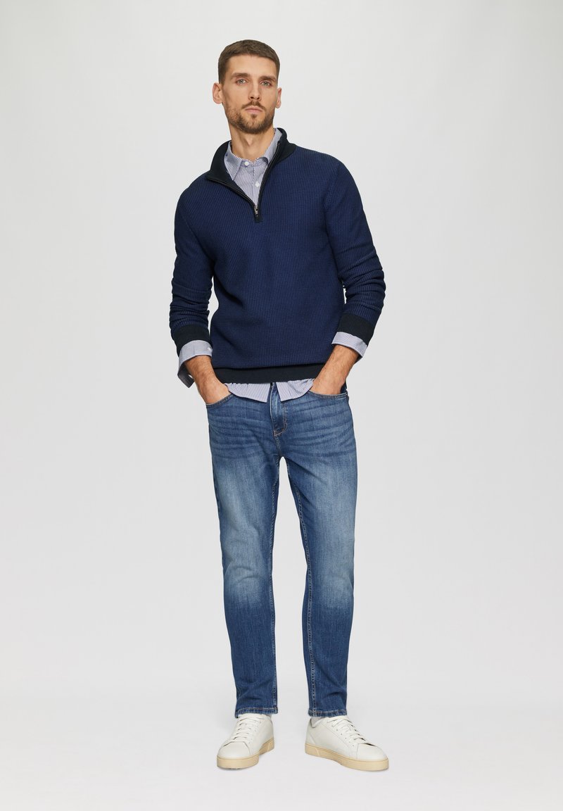 Marineblaue Strick-Pullover mit halbem Reißverschluss über einem hell gestreiften Hemd, kombiniert mit hellblauen Jeans und weißen Turnschuhen mit beigen Sohlen. Einfaches Design.