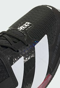 Sort sportsko med en textureret mesh overdel, blanke detaljer, stort hvidt logo og rød sål med præget tekst.