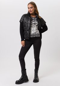 Schwarz gepolsterte Weste mit hohem Kragen über einem schwarz-weißen gemusterten Pullover, kombiniert mit schwarzen Slim-Fit-Hosen und kniehohen schwarzen Stiefeln.