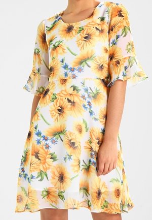 Žena nosící bílé šaty s velkými žlutými vzory slunečnic a malými modrými květy, s 3/4 zvonovými rukávy a kulatým výstřihem.